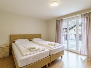 A double bed with white linens and a wooden headboard in a room with a wooden floor and a large window with curtains. - Haus Astoria by Interhome (Saas-Grund)
