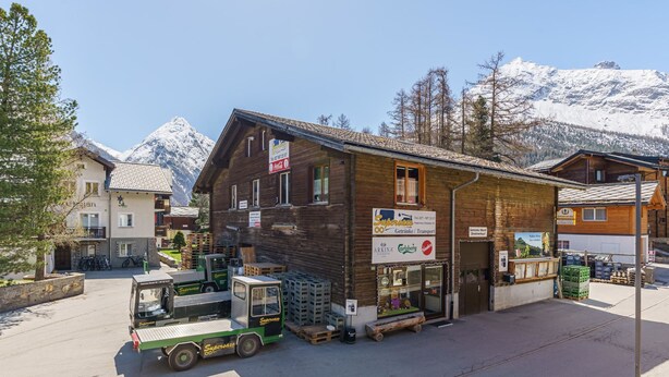 Una cabaña de montaña con fachada de madera, una camioneta estacionada al frente y una vista despejada de montañas nevadas al fondo.