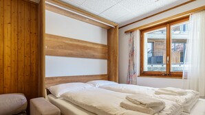 Una habitación con cabecera de madera, cama con ropa blanca y toallas, un banco y una ventana con cortinas.