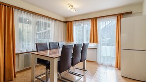 A modern dining area with a wooden table and chairs, large windows with curtains, and a white refrigerator. - Haus City by Interhome (Saas-Grund)