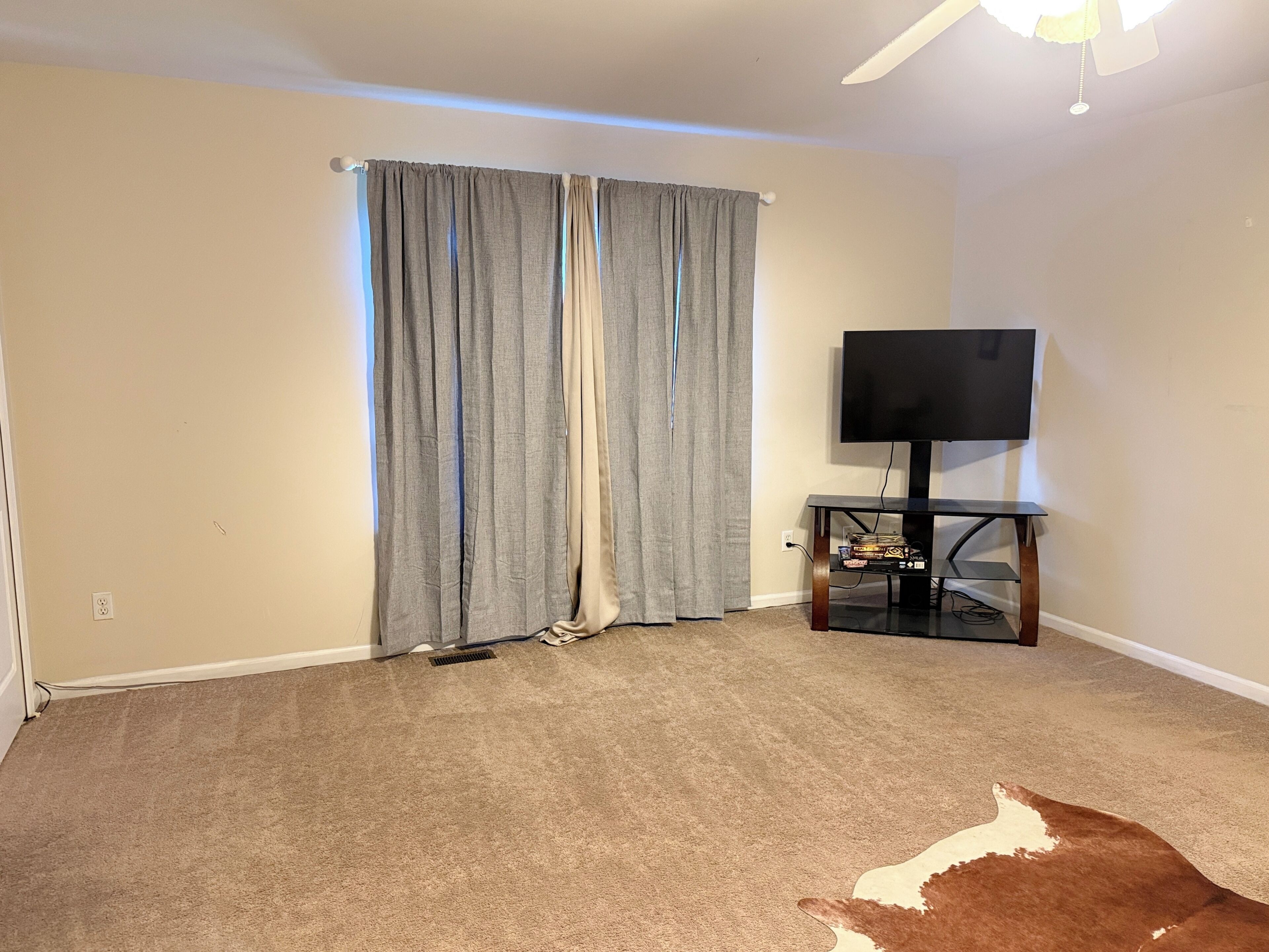 Una habitación con alfombra color beige, un televisor sobre un mueble bajo y una ventana con cortinas grises.