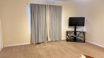 Une pièce avec un tapis beige, un téléviseur posé sur un meuble et une fenêtre avec des rideaux gris.