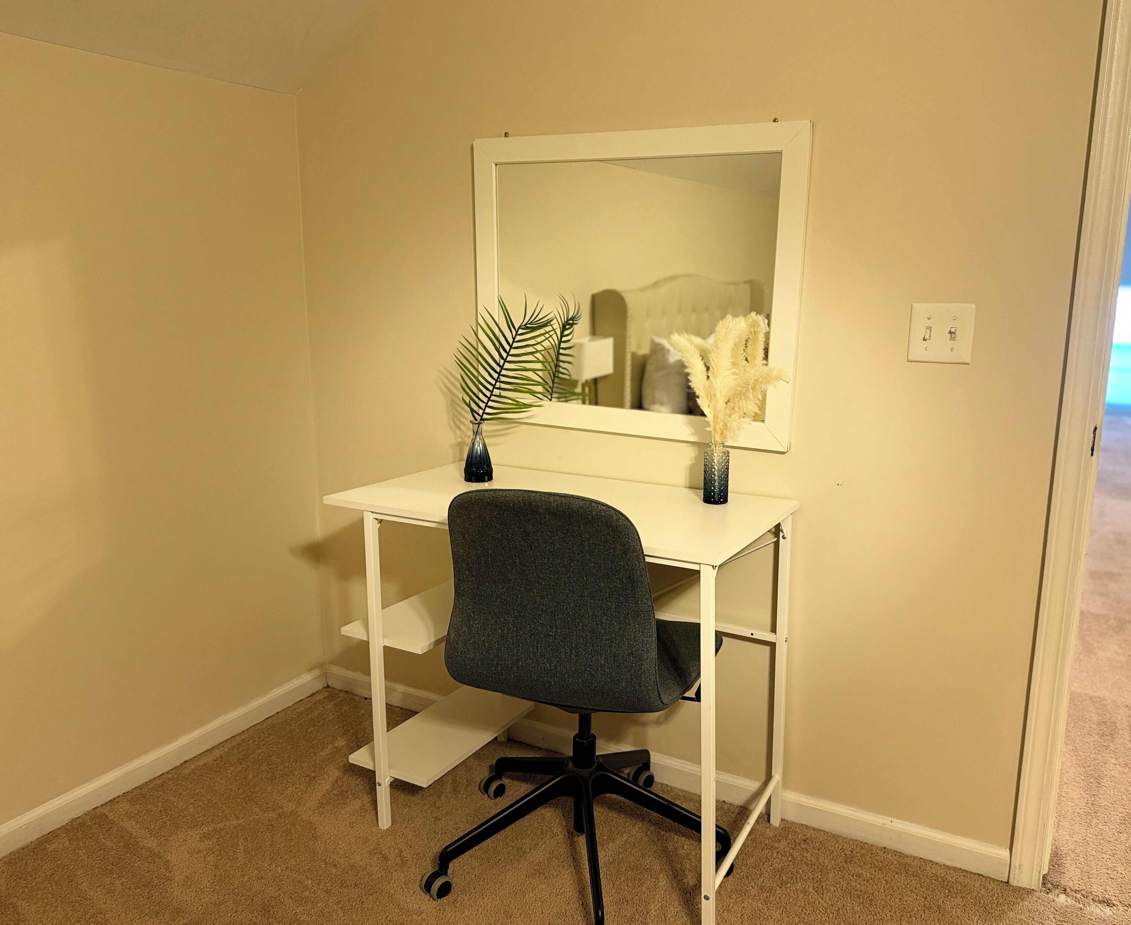 Un bureau blanc avec un miroir, un fauteuil de bureau foncé et une petite plante dans un vase.