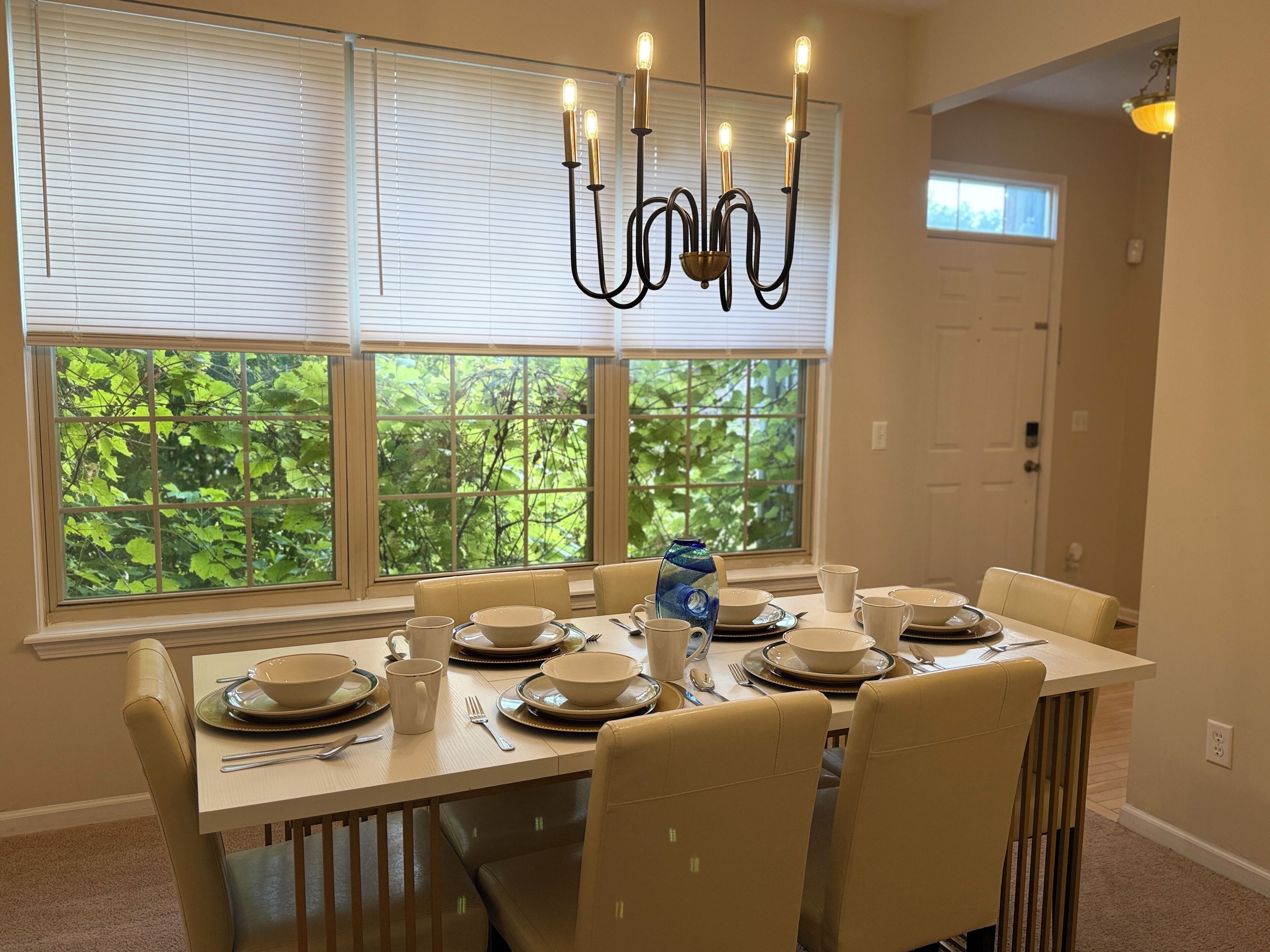 Un coin repas avec une table dressée pour quatre personnes, un lustre et une vue sur la verdure par les fenêtres.