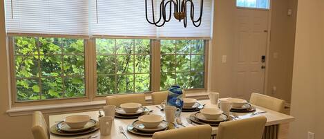 A dining area with a table set for four, a chandelier, and a view of greenery through the windows.