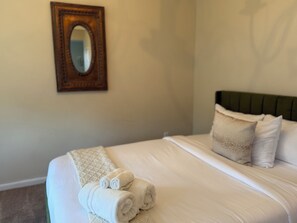 A neatly made bed with white linens and rolled towels, a green upholstered headboard, and a mirror on the wall.