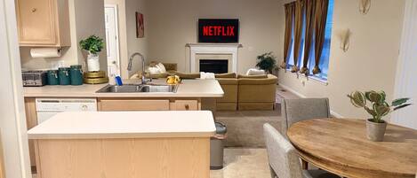 A modern kitchen with a built-in dishwasher, a dining area with a wooden table and chairs, a sofa, and a fireplace.