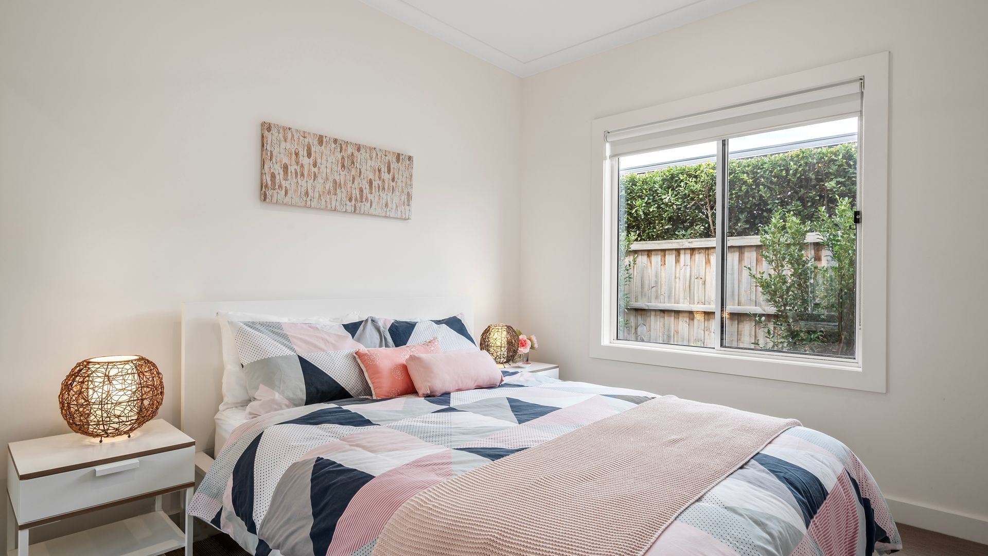 A bedroom with a bed, bedside tables, a window with a view of greenery, and a wall-mounted artwork.