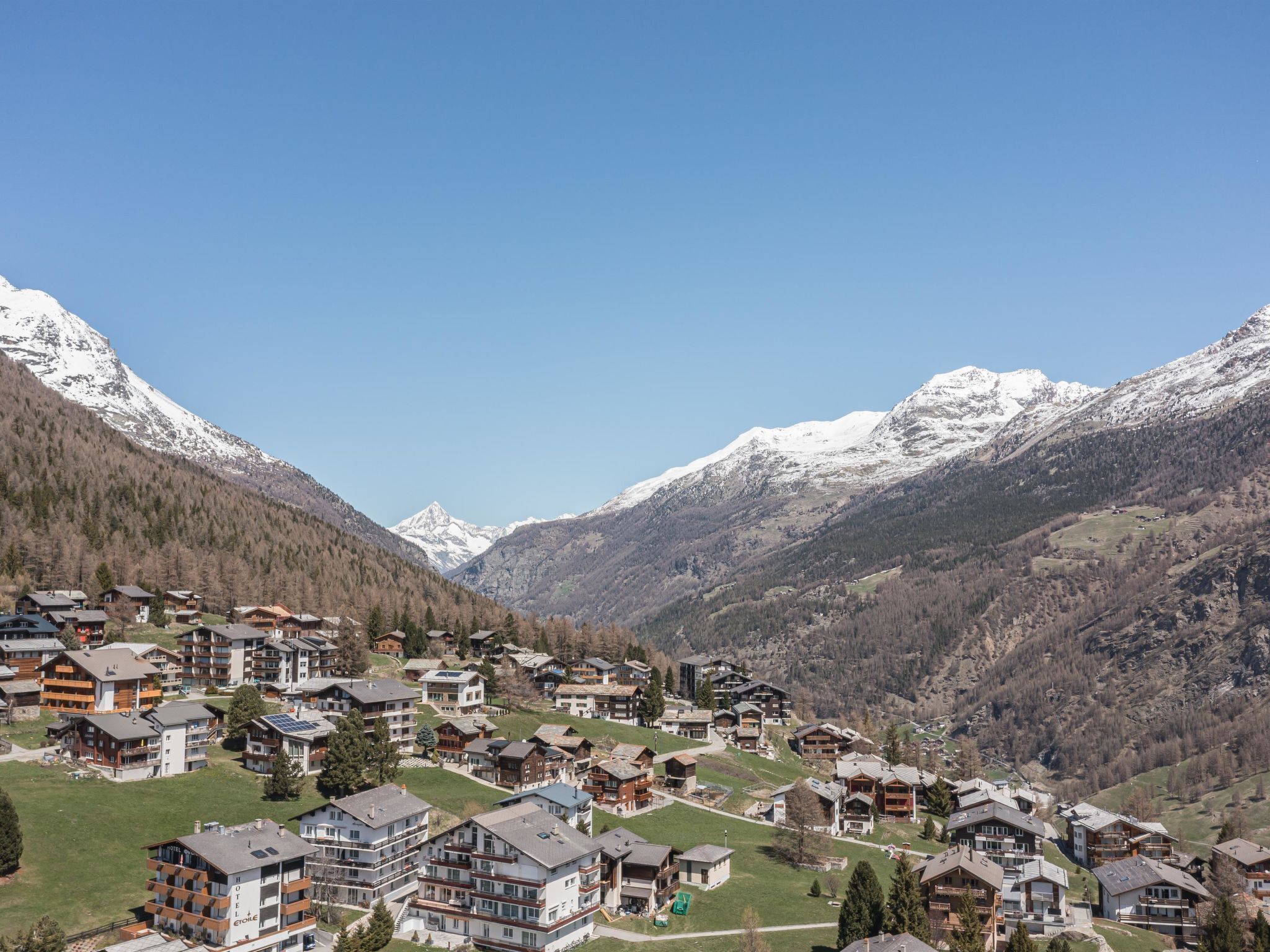 Ein Bergdorf mit Chalets, strahlend blauem Himmel und schneebedeckten Bergen im Hintergrund.