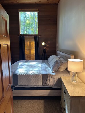 Une pièce en bois avec un lit, une table de chevet, une lampe et une fenêtre avec des rideaux.