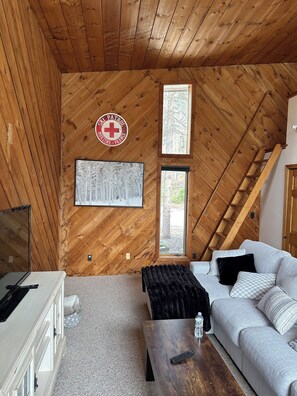 Ein gemütliches Zimmer mit Holzwänden, einem Schild der Skiwacht, einem gerahmten Bild, einem Sofa, einem Couchtisch und einem Fernseher.
