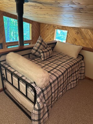 Ein Bett mit kariertem Bettbezug und Kissen, eine flauschige, weiße Decke und Blick auf Bäume durch ein Fenster.