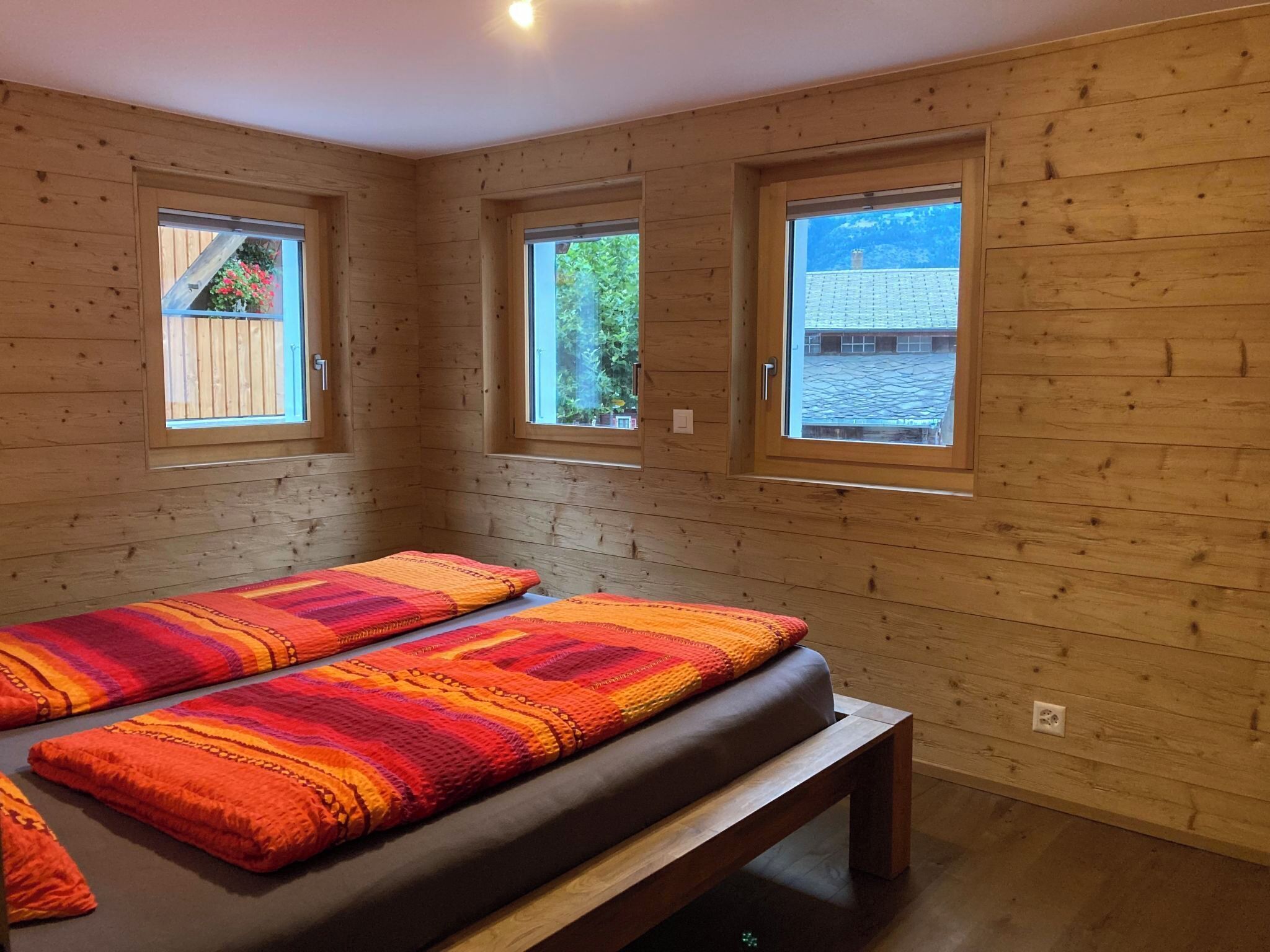 Una habitación con paredes de madera, una cama con un edredón colorido y tres ventanas.