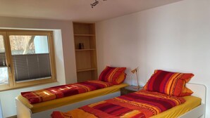 Een slaapkamer met twee enkele bedden, een raam met zonwering, een houten plank en een bedlampje.