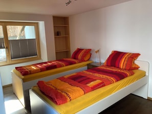 Ein Schlafzimmer mit zwei Einzelbetten, einem Fenster mit Jalousien, einem Holzregal und einer Nachttischlampe.