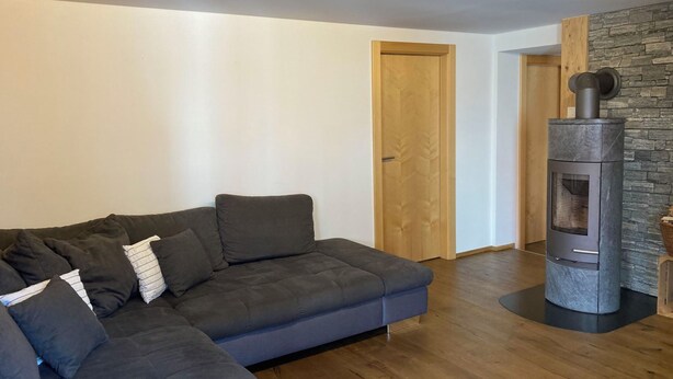 Een moderne woonkamer met een donkergrijze hoekbank, een stenen muur en een houtkachel.