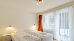 A hotel room with two beds, a nightstand, a wall-mounted light, and a window with sheer curtains. - Haus City by Interhome (Saas-Grund)