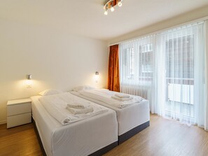 A hotel room with two beds, a nightstand, a wall-mounted light, and a window with sheer curtains. - Haus City by Interhome (Saas-Grund)
