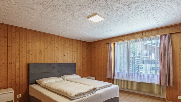 Ein Schlafzimmer mit einer holzverkleideten Wand, einem großen Bett mit weißer Bettwäsche, einem Nachttisch, einem Fenster mit Vorhängen und einer Decke mit Rastermuster.