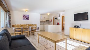 Un salon moderne avec un canapé, une table à manger et des chaises. Il y a un coin cuisine avec des placards et un four à micro-ondes. Un téléviseur est fixé au mur.