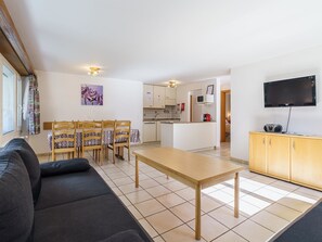 Ein modernes Wohnzimmer mit einer Couch, einem Esstisch und Stühlen. Es gibt eine Küchenzeile mit Schränken und einer Mikrowelle. Ein Fernseher ist an der Wand befestigt.