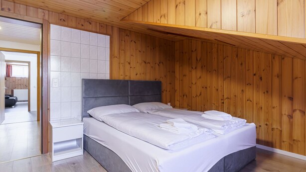 A bedroom with a wooden ceiling, a bed with white bedding, and a view of a bathroom through an open door. - Haus Slalom by Interhome (Saas-Fee)
