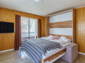 Ein modernes Schlafzimmer mit Holzboden, einem Bett mit gemustertem Überwurf, einem Flachbildfernseher und einem großen Fenster mit Vorhängen.