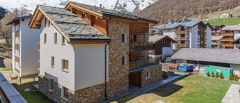 Un immeuble à plusieurs étages, doté d’une façade en pierre et de détails en bois, se dressant sur fond de montagnes enneigées.