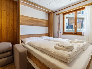 A room with a wooden headboard, a bed with white linens and towels, a bench, and a window with curtains. - Haus Slalom by Interhome (Saas-Fee)