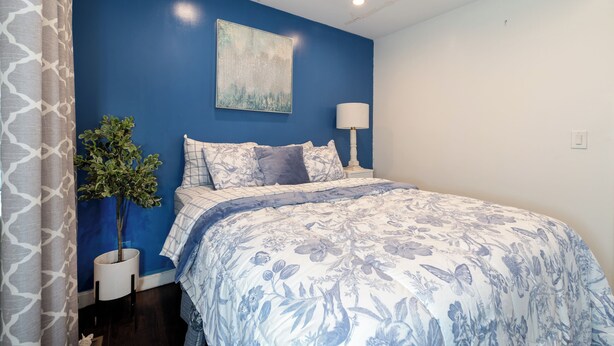 A bedroom with a blue accent wall, a bed with floral bedding, a potted plant, and a bedside lamp. - Cozy getaway (Oakland)