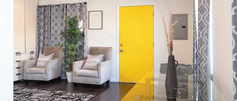 A modern living room with two armchairs, a glass dining table, a yellow door, and a patterned curtain.