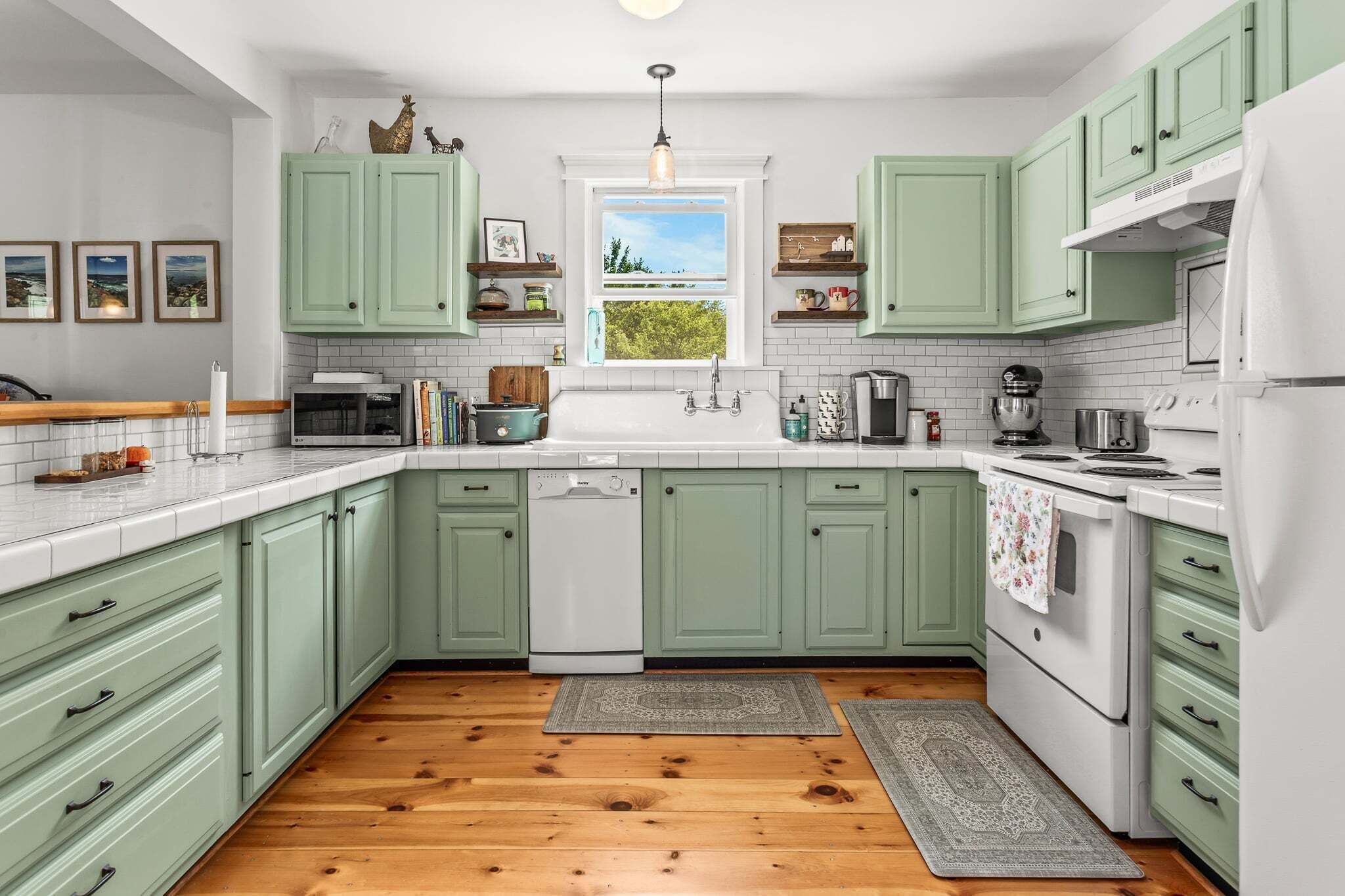 A kitchen with green cabinets, a white countertop, a dishwasher, a sink, and a window with a view of trees.
