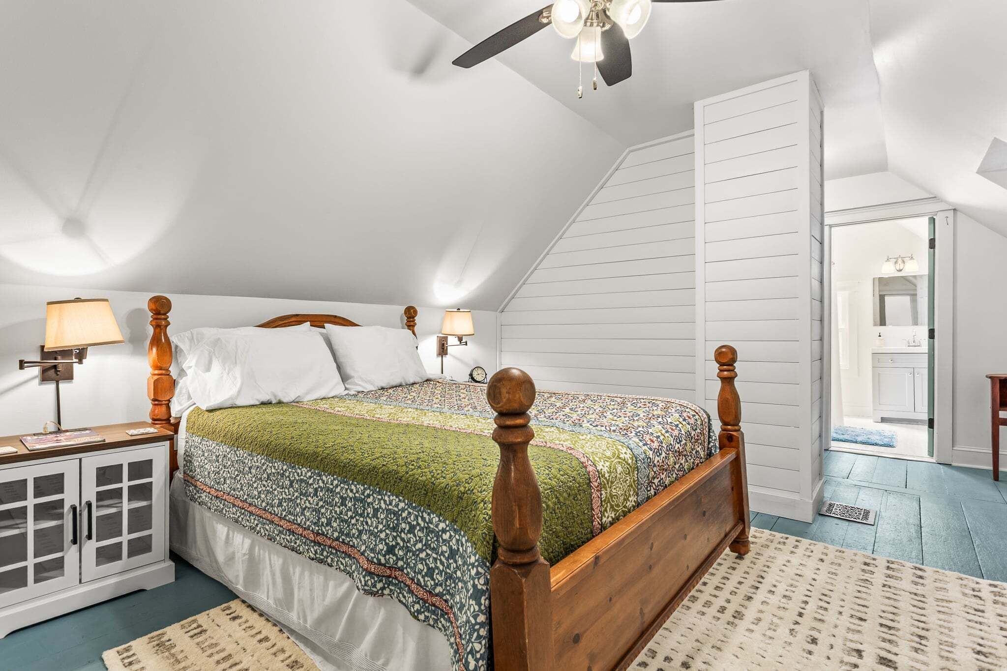 A bedroom with a wooden four-poster bed, a nightstand with a lamp, a wardrobe, and a ceiling fan.