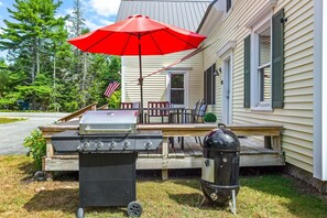 Ein Grillset mit Gas- und Holzkohlegrill ist auf einer Holzterrasse mit einem roten Sonnenschirm aufgebaut.