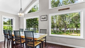 A dining room with a large table and chairs, large windows with a view of trees and a lake, and a sign on the wall. - Patten Pond Retreat - NEW! Waterfront (Surry)