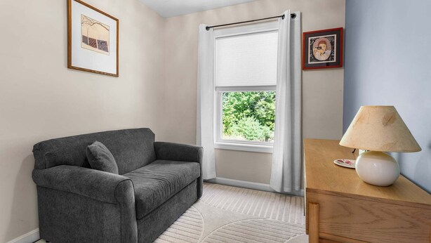 A room with a grey sofa, a wooden side table with a lamp, a window with white curtains, and a framed picture on the wall. - Patten Pond Retreat - NEW! Waterfront (Surry)