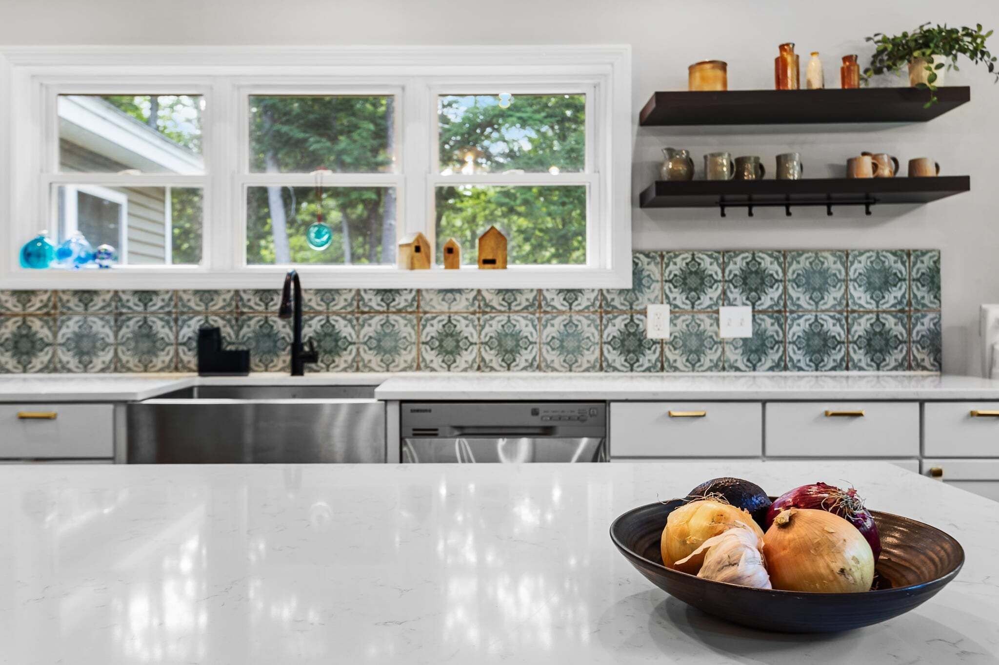 A kitchen with a white countertop, a stainless steel sink, a dishwasher, and a window with a view of trees. There are decorative items on the windowsill and a bowl of vegetables on the counter.