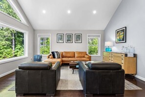Un salon spacieux avec une grande fenêtre, un canapé en cuir marron, deux poufs en cuir noir, un buffet en bois et des tableaux encadrés accrochés au mur.