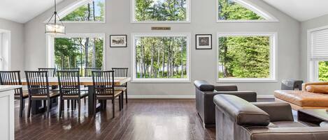 A spacious room with a dining area, a seating area with a sofa, and large windows offering views of trees and a lake.