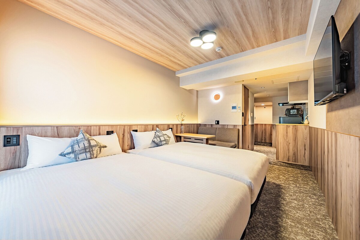 Ein Hotelzimmer mit zwei Betten, einem wandmontierten Flachbildfernseher, einem kleinen Tisch und einer Küchenzeile.