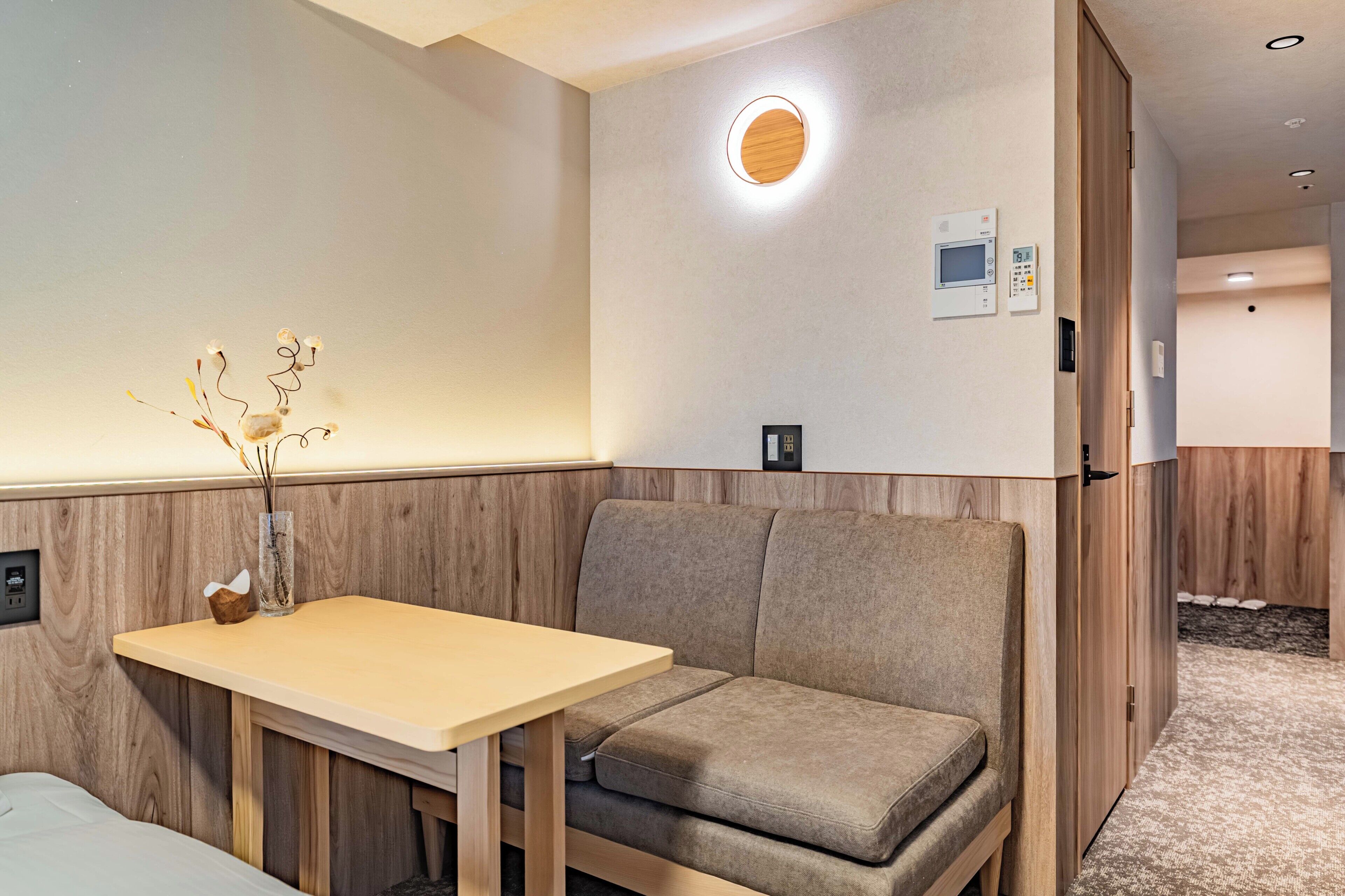 Camera d'hotel con parete decorata in legno, un piccolo tavolo con un vaso di piante secche, una panca imbottita, una TV a muro e un termostato.