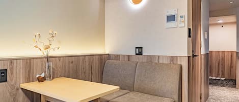 A hotel room with a wooden accent wall, a small table with a vase of dried plants, a cushioned bench, a wall-mounted TV, and a thermostat.