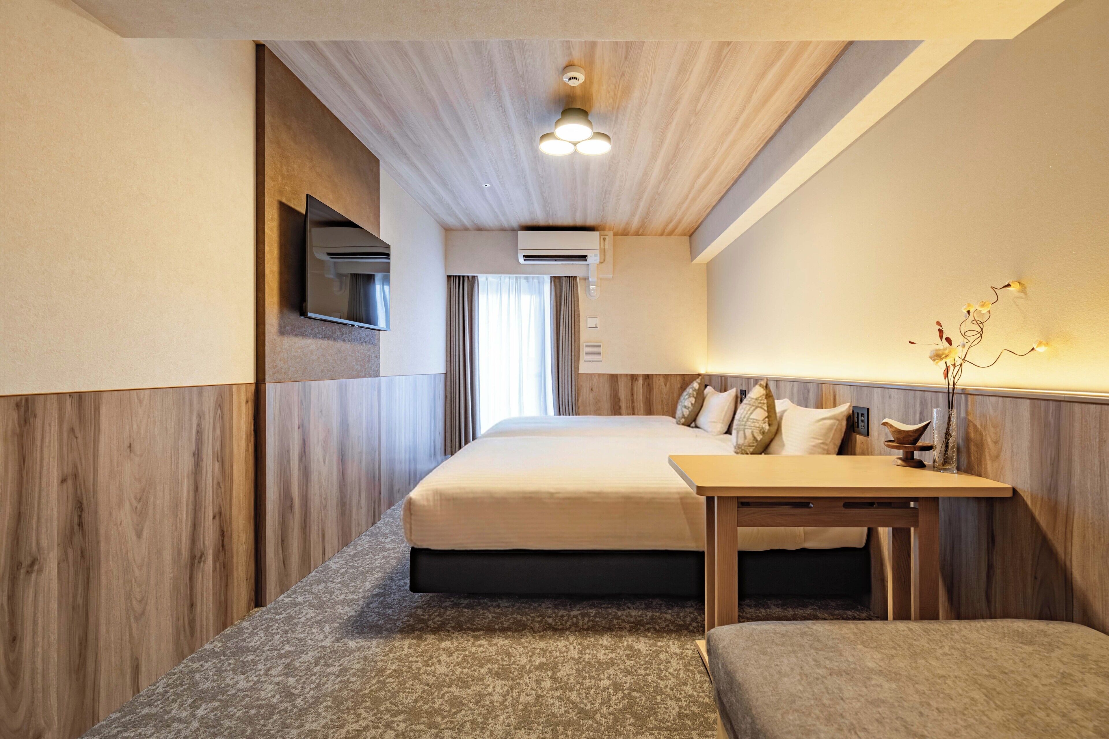 A hotel room with a bed, a desk, a mirror, an air conditioning unit, and a window with curtains.