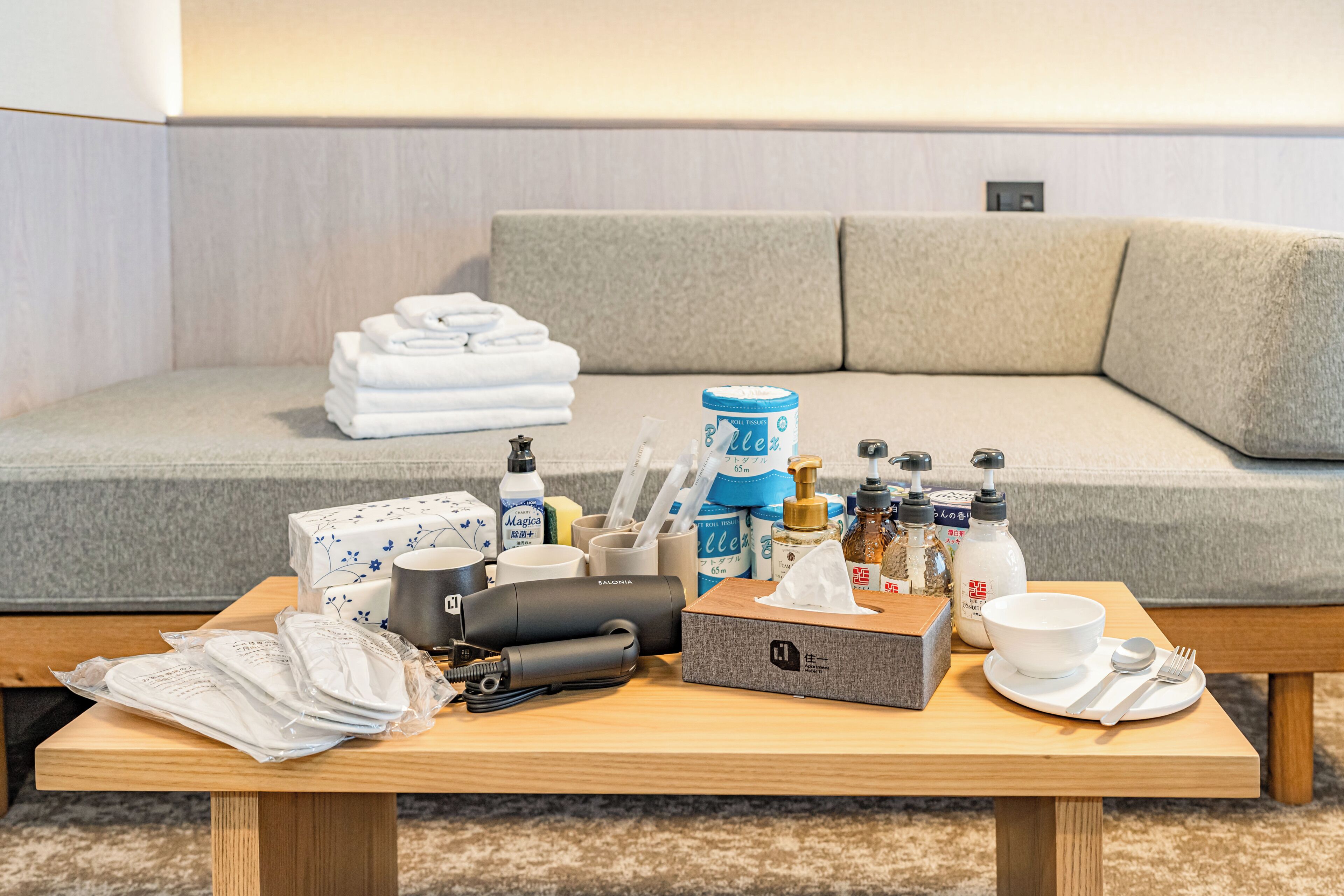 A neatly arranged table with toiletries, a tissue box, a mug, and a plate with cutlery in front of a sofa.