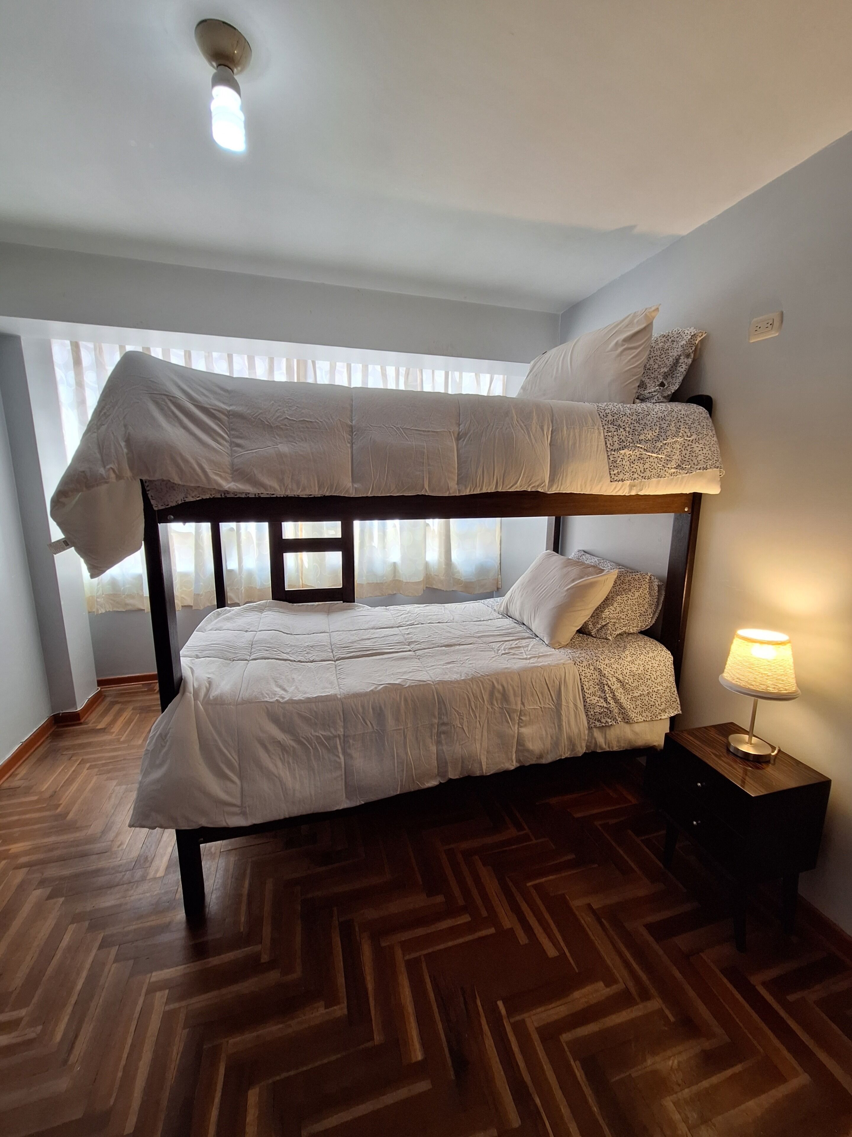 A bunk bed with a wooden frame and white bedding, a bedside table with a lamp, and a herringbone-patterned wooden floor.