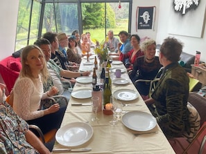 A group of people seated around a table with plates, glasses, and bottles, in a room with a window and a wall with a framed picture.