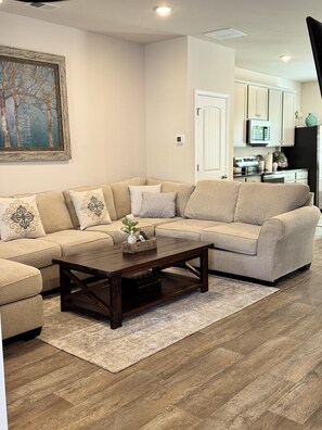 A living room with a beige sectional sofa, a wooden coffee table, and a rug on a wooden floor.