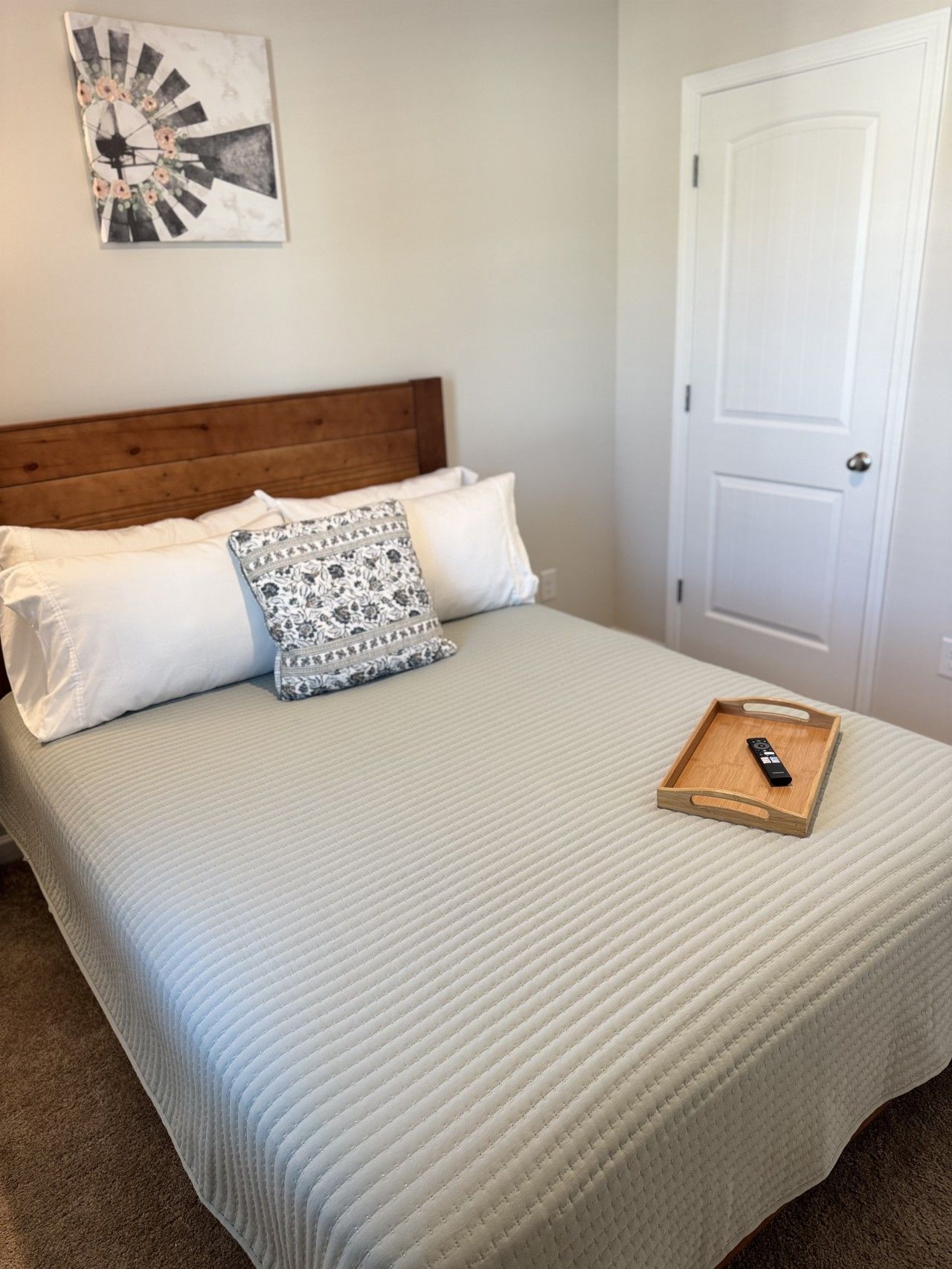 Un lit bien fait, avec une tête de lit en bois, deux oreillers, un coussin décoratif et un plateau sur lequel se trouve une télécommande, le tout posé sur une couverture au motif texturé.
