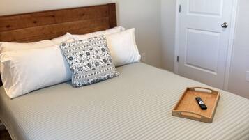 Un lit bien fait, avec une tête de lit en bois, deux oreillers, un coussin décoratif et un plateau sur lequel se trouve une télécommande, le tout posé sur une couverture au motif texturé.