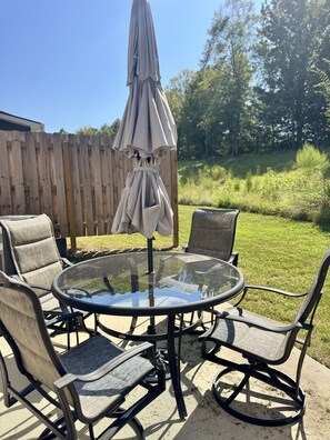 A round glass table with four chairs, an umbrella, and a wooden fence in the background.